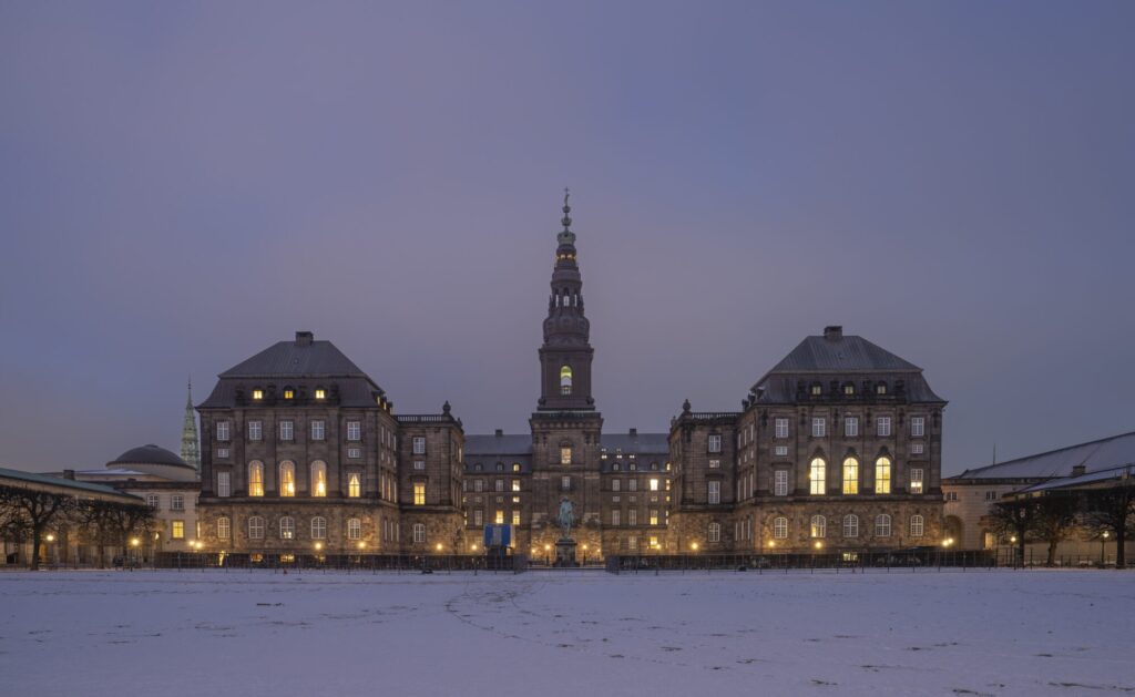 Christiansborg set fra Ridebanen en vinteraften med sne på pladsen og lys i vinduerne.